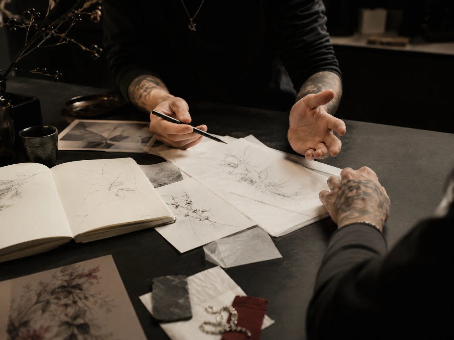 Two pairs of hands above layered botanical sketches, tracing paper, and material samples on a dark consultation table.
